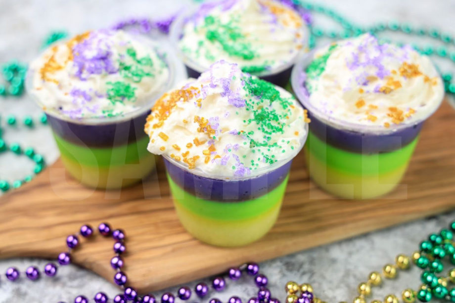 The Mardi Gras Layered Jello Shots comes in a clear jello shot cup with a gray striped napkin on a marble backdrop.
