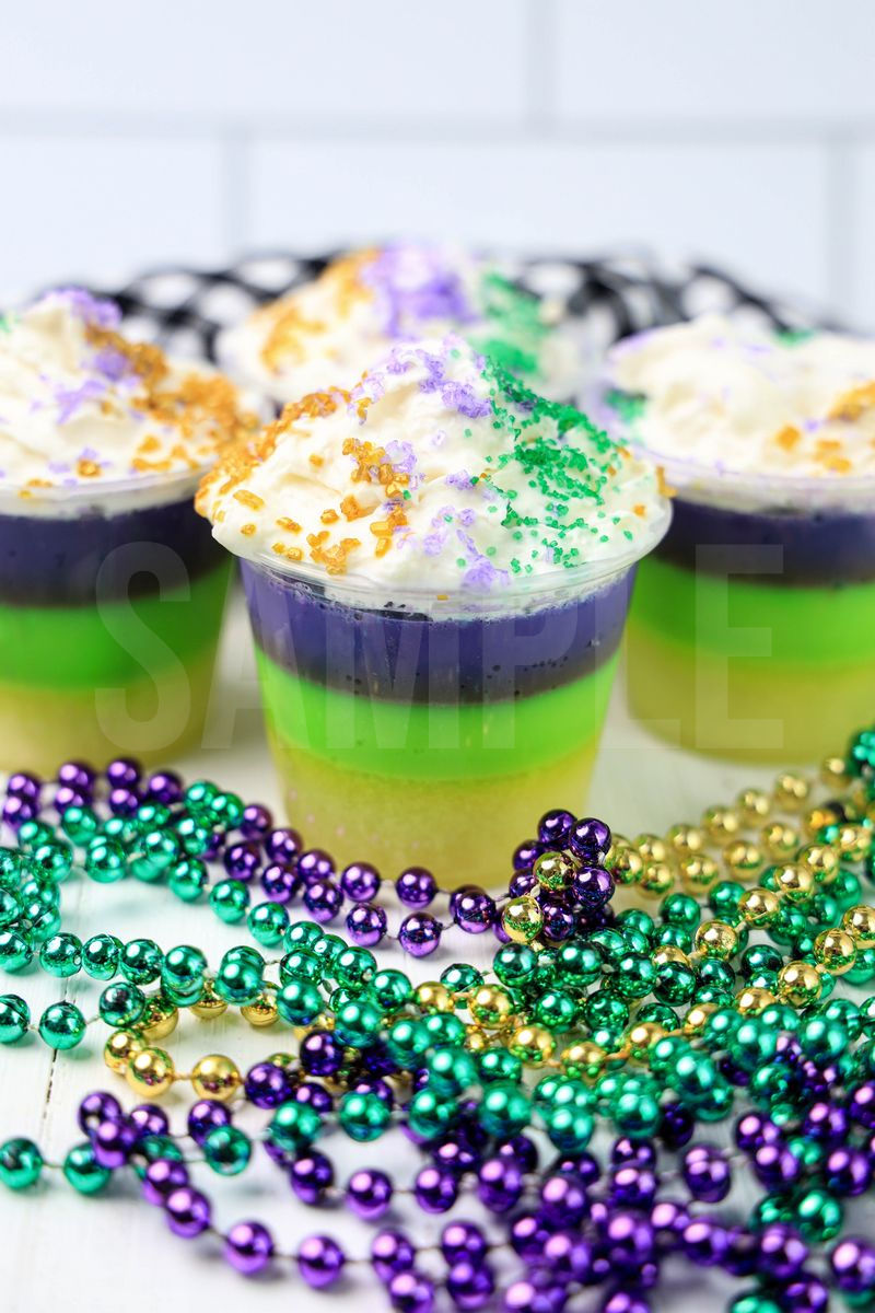 The Mardi Gras Layered Jello Shots comes in a clear jello shot cup with a plaid napkin on a white wood backdrop.
