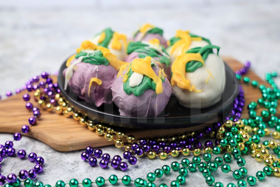 The Mardi Gras Oreo Balls comes on a black plate on a olive wood cutting board on a marble backdrop.