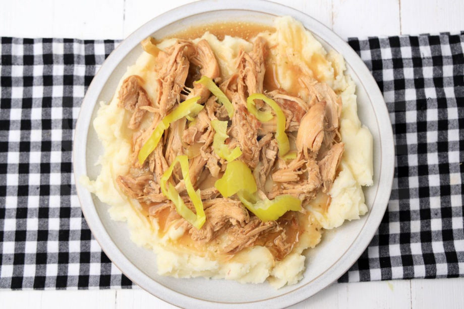 The Slow Cooker Mississippi Chicken comes on a stone plate with a plaid napkin on a white wood backdrop