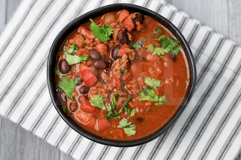 The Slow Cooker Beef Chili comes in a black bowl with a white striped napkin on a gray wood backdrop.