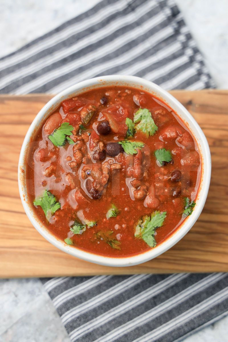 The Slow Cooker Beef Chili comes in a white bowl with a gray striped napkin on a marble backdrop.