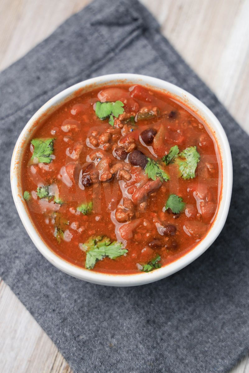 The Slow Cooker Beef Chili comes in a white bowl with a denim napkin on a rustic wood backdrop.