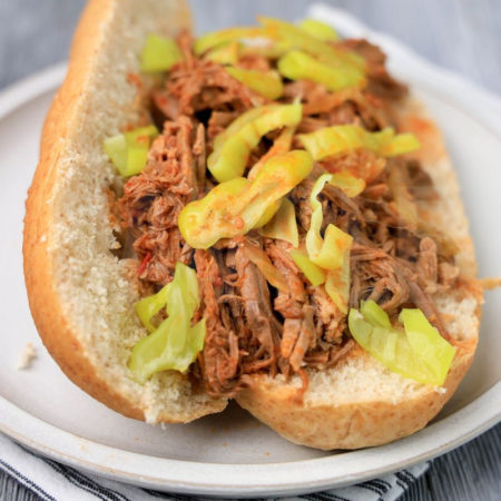 The Slow Cooker Italian Beef Sandwiches comes on a stone plate with a white striped napkin on a gray wood backdrop.