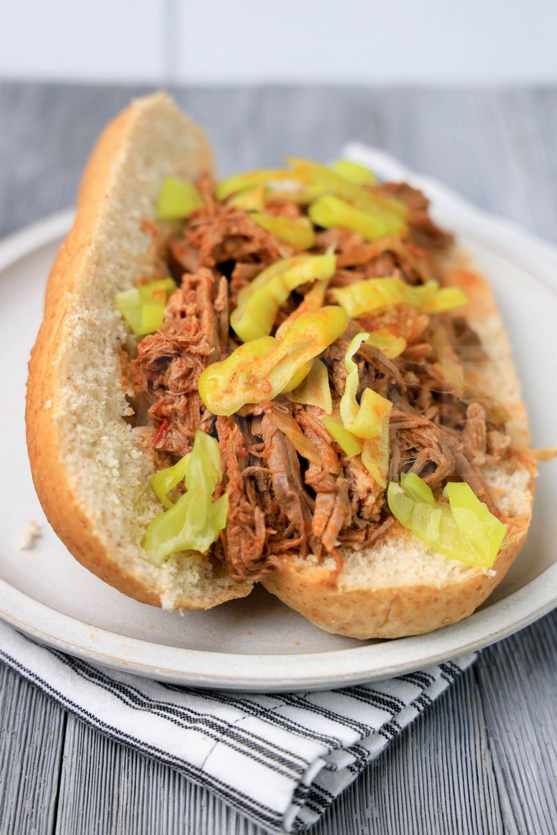 The Slow Cooker Italian Beef Sandwiches comes on a stone plate with a white striped napkin on a gray wood backdrop.