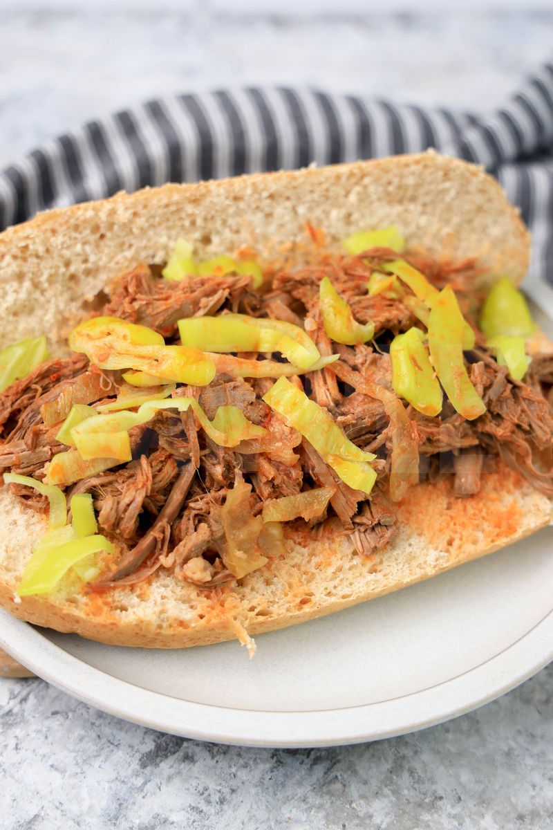 The Slow Cooker Italian Beef Sandwiches comes on a stone plate with a gray striped napkin on a marble backdrop.