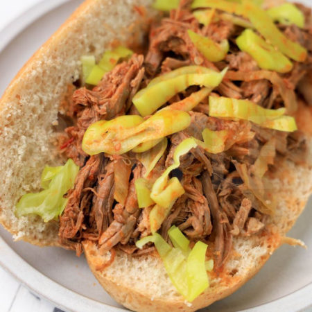 The Slow Cooker Italian Beef Sandwiches comes on a stone plate with a plaid napkin on a white wood backdrop.