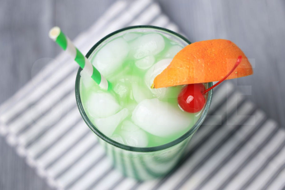 The Green Drunken Leprechaun comes in a clear glass with a white striped napkin on a gray wood backdrop.