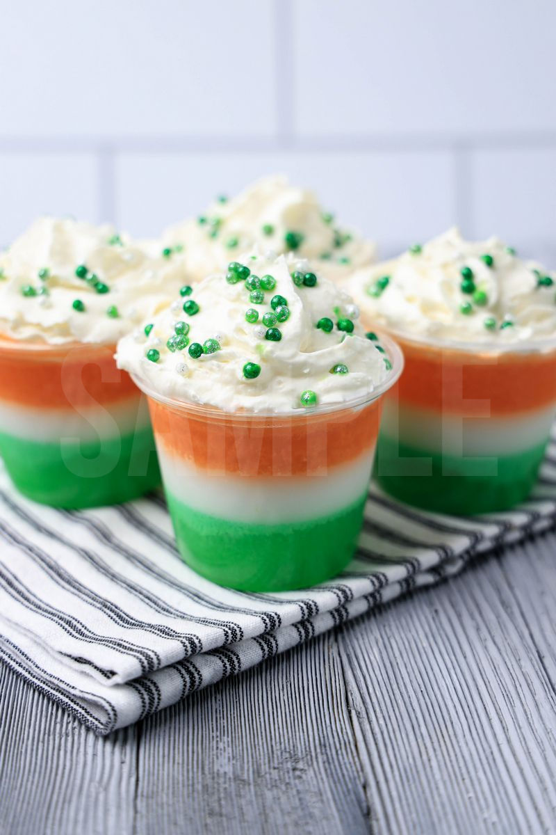 The Irish Flag Jello Shots come in a clear plastic cup with a white striped napkin on a gray wood backdrop.