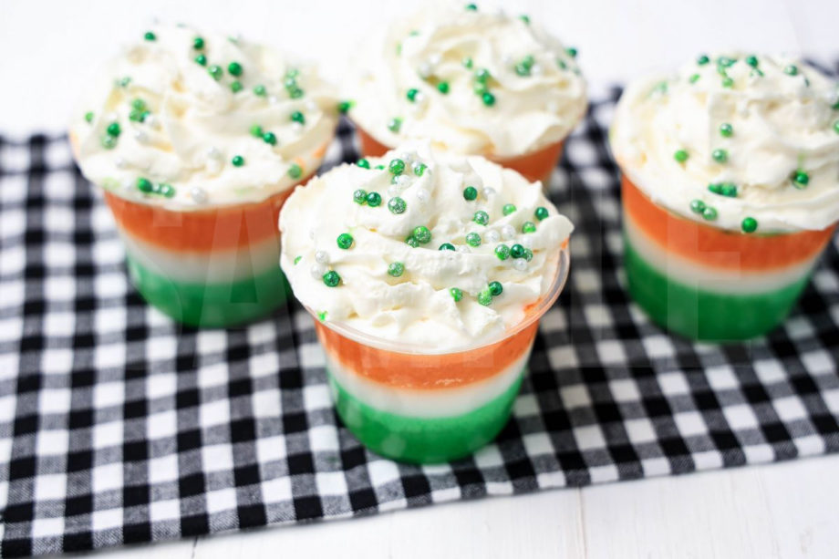 The Irish Flag Jello Shots come in a clear plastic cup with a plaid napkin on a white wood backdrop.