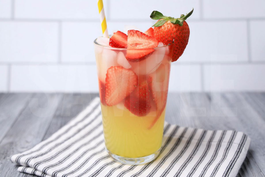 The Limoncello Strawberry Lemonade comes in a clear glass with a white striped napkin on a gray wood backdrop.