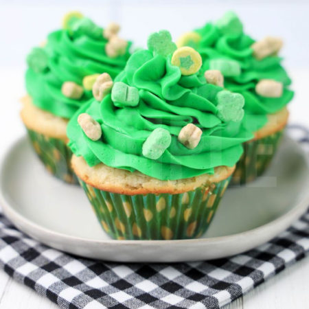 The Lucky Charms Cupcakes comes on a white plate on a plaid napkin with a white wood backdrop.