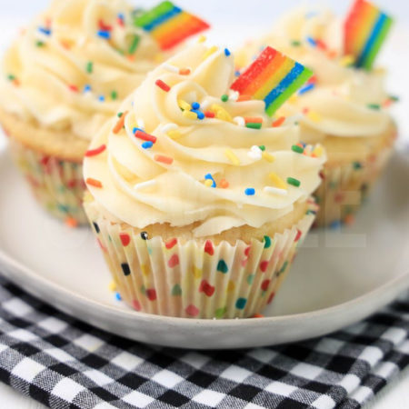 The Rainbow Cupcakes comes on a white plate on a gray striped napkin with a marble backdrop.