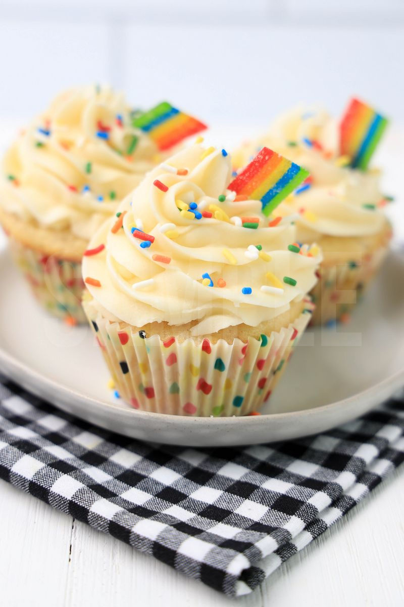 The Rainbow Cupcakes comes on a white plate on a gray striped napkin with a marble backdrop.