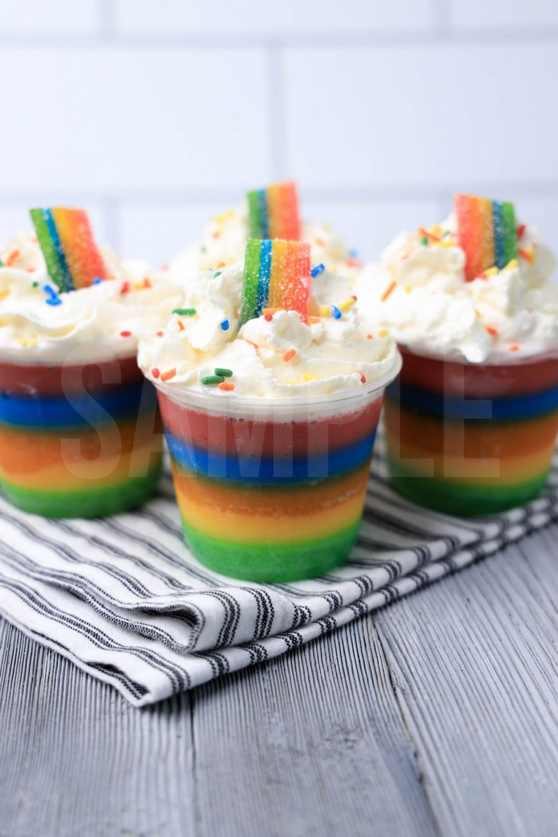 The Rainbow Jello Shots come in a clear plastic cup with a white striped napkin on a gray wood backdrop.