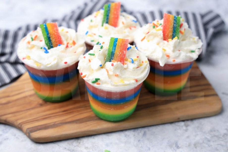 The Rainbow Jello Shots come in a clear plastic cup with a gray striped napkin on a marble backdrop.