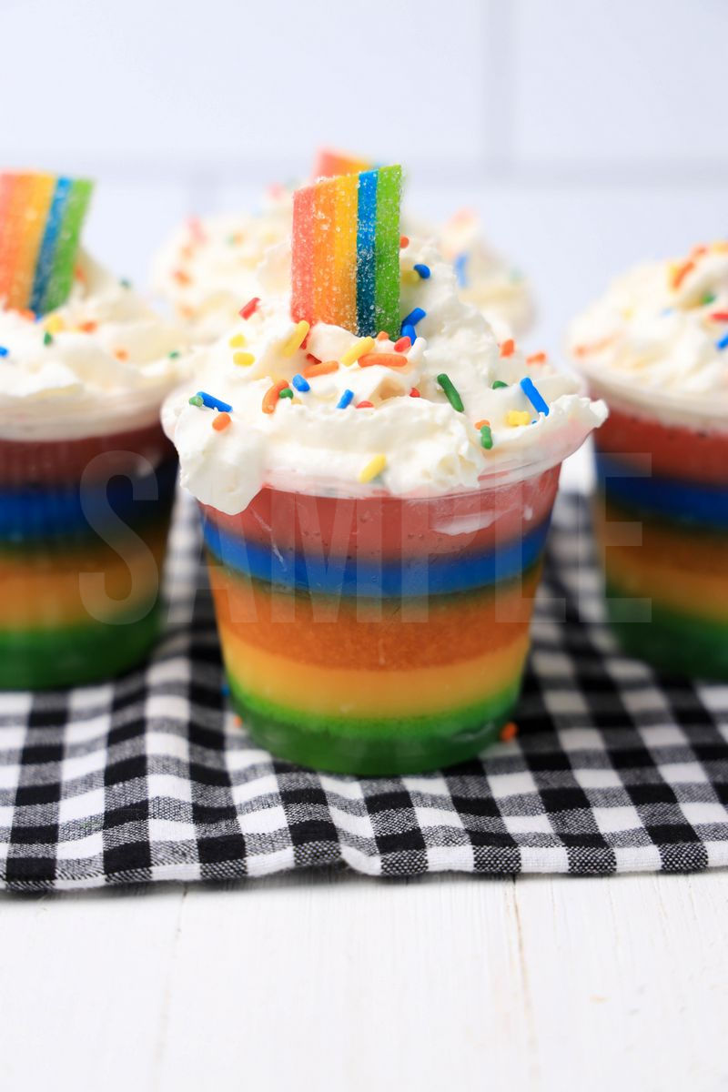 The Rainbow Jello Shots come in a clear plastic cup with a plaid napkin on a white wood backdrop.
