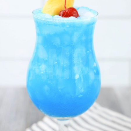 The Blue Hawaii Cocktail comes in a hurricane glass with a white striped napkin on a gray wood backdrop.