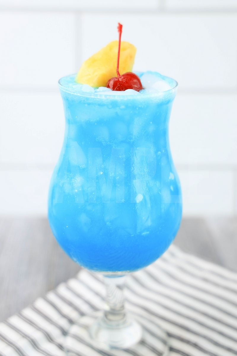 The Blue Hawaii Cocktail comes in a hurricane glass with a white striped napkin on a gray wood backdrop.