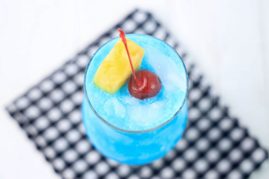 The Blue Hawaii Cocktail comes in a hurricane glass with a plaid napkin on a white wood backdrop.