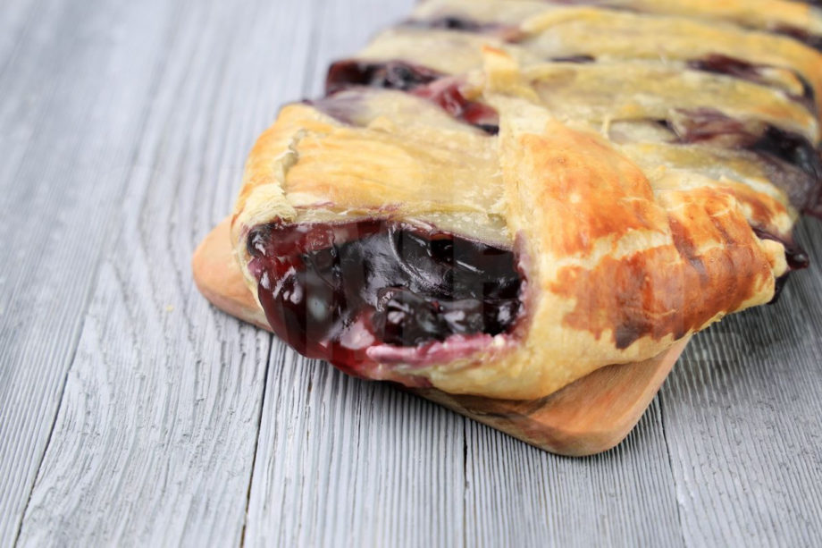 The Blueberry Puff Pastry Braid comes on a olive wood board on a gray wood backdrop.