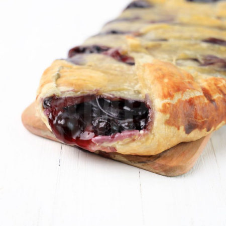 The Blueberry Puff Pastry Braid comes on a olive wood board on a white wood backdrop.