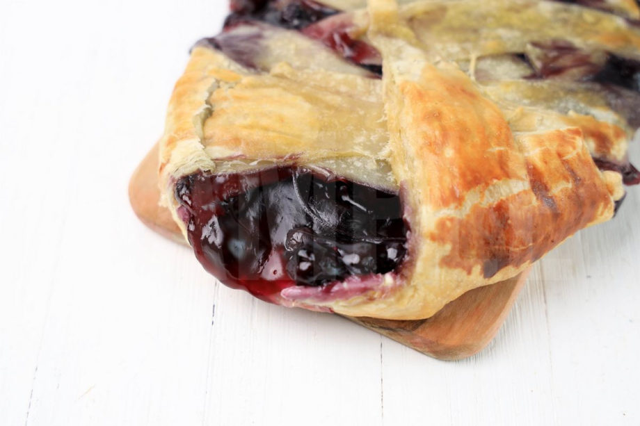 The Blueberry Puff Pastry Braid comes on a olive wood board on a white wood backdrop.