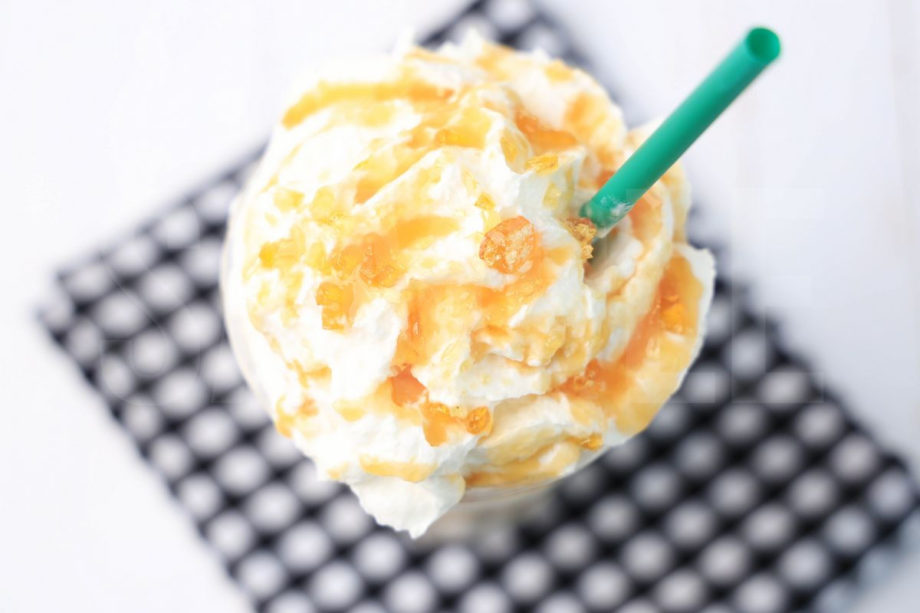 The Caramel Ribbon Crunch Crème Frappuccino comes in a venti cup with a plaid napkin on a white wood backdrop.