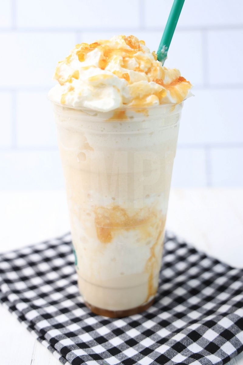 The Caramel Ribbon Crunch Crème Frappuccino comes in a venti cup with a plaid napkin on a white wood backdrop.