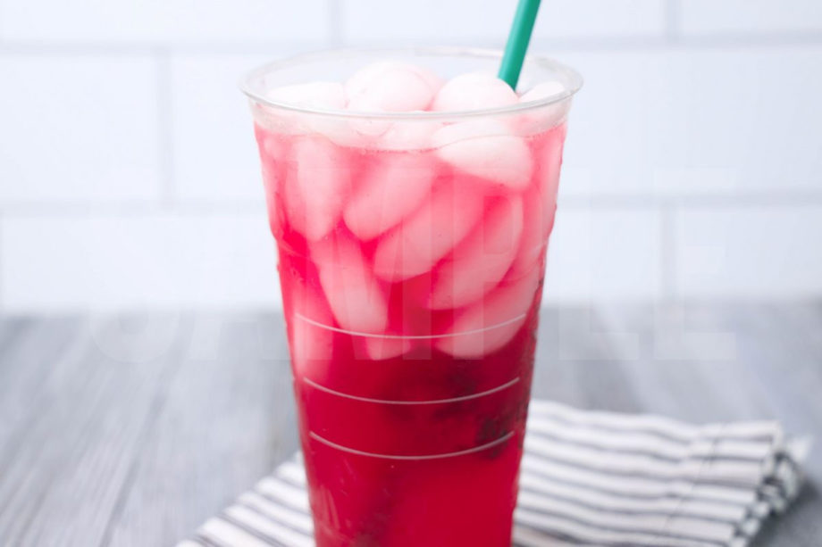 The Mango Dragonfruit Refresher comes in a venti cup with a white striped napkin on a gray wood backdrop.