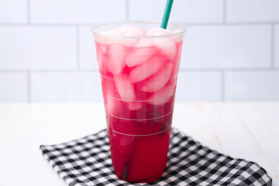 The Mango Dragonfruit Refresher comes in a venti cup with a plaid napkin on a white wood backdrop.