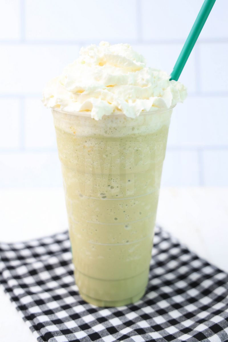 The Matcha Crème Frappuccino comes in a venti cup with a plaid napkin on a white wood backdrop.