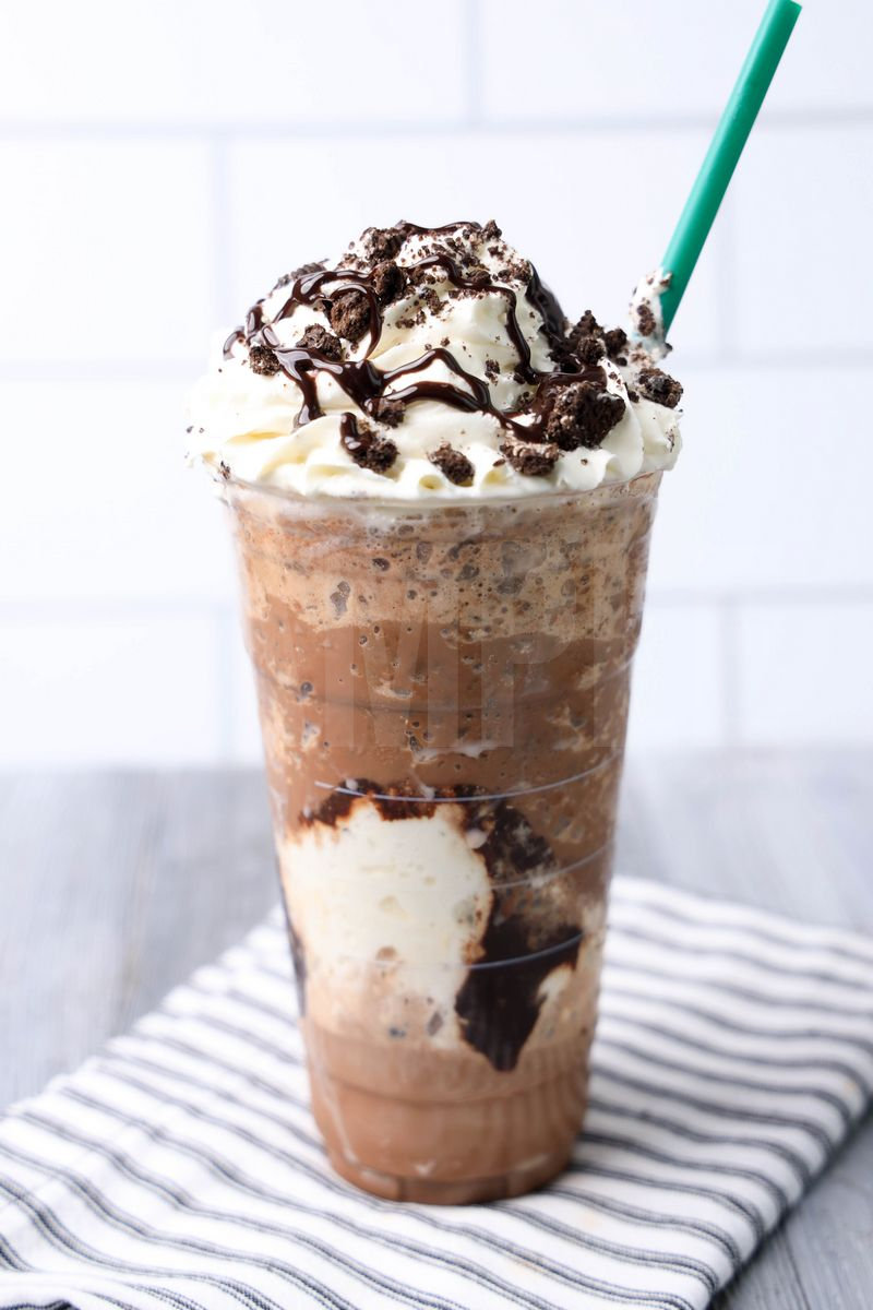 The Mocha Cookie Crumble Frappuccino comes in a venti cup with a white striped napkin on a gray wood backdrop.