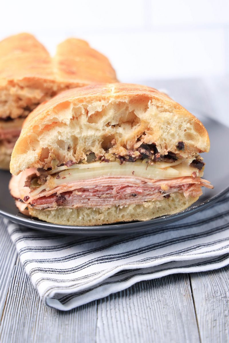 The Ciabatta Muffuletta comes on a gray plate with white striped napkin on a gray wood backdrop.