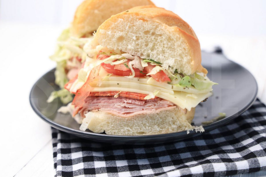 The Italian Grinder sandwich comes on a gray plate with plaid napkin on a white wood backdrop.
