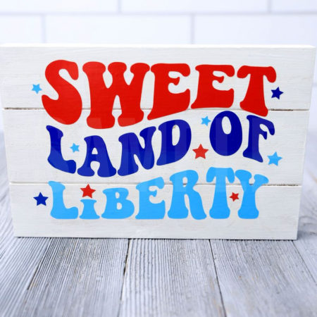 The Sweet Land Of Liberty Farmhouse Wood Cricut Craft comes pictured on a gray wood backdrop.