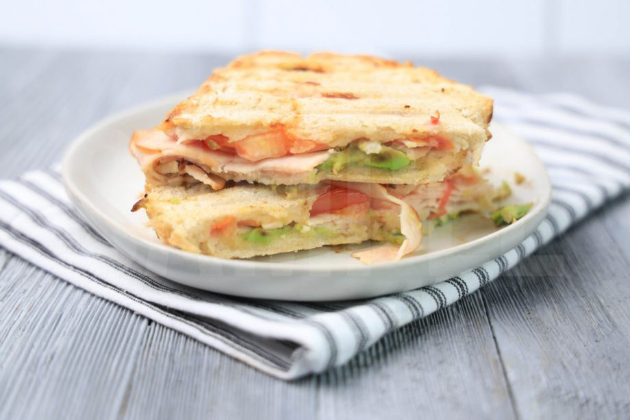 The Turkey Avocado Panini comes on a white plate with a white striped napkin on a gray wood backdrop.