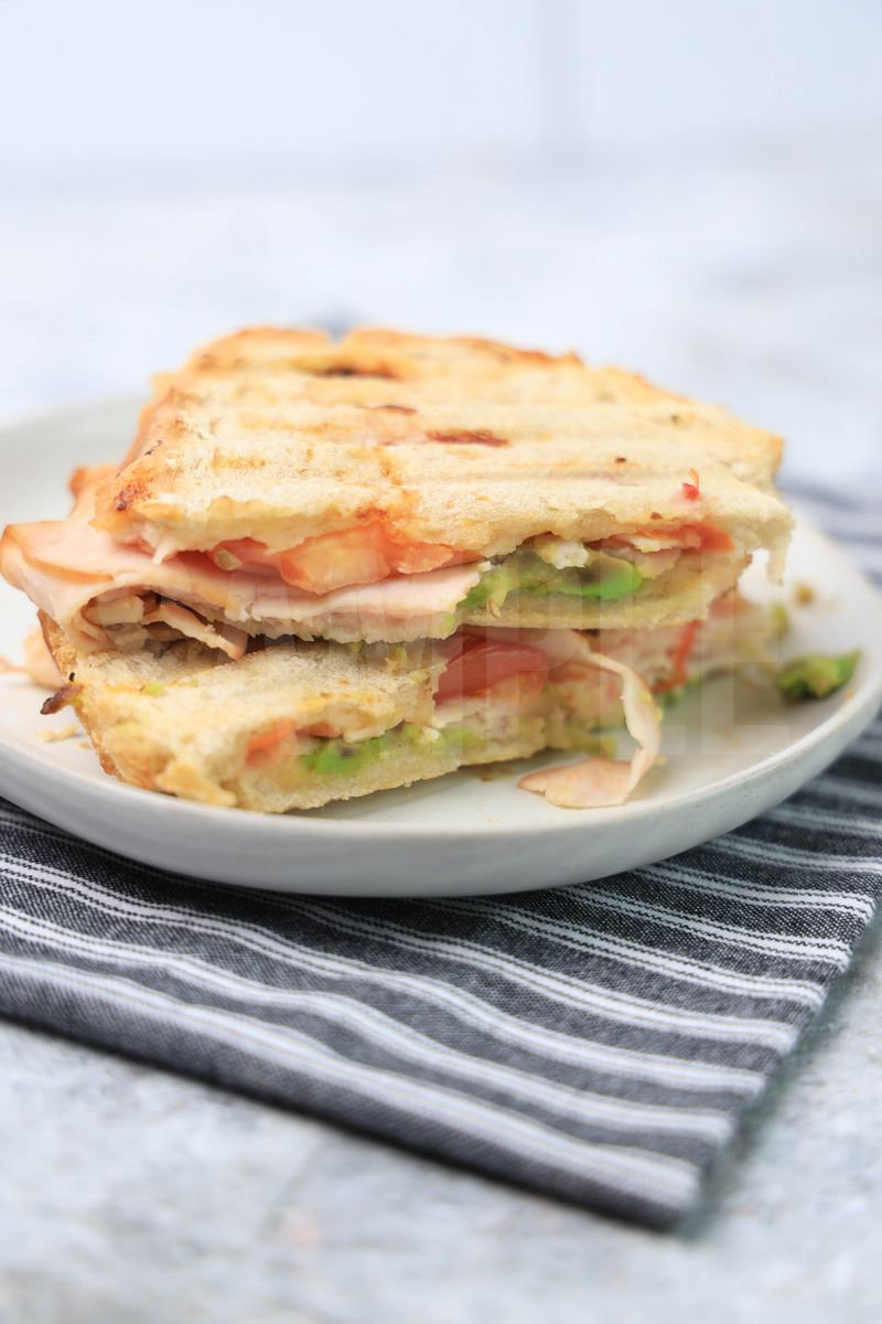 The Turkey Avocado Panini comes on a white plate with a gray striped napkin on a marble backdrop.