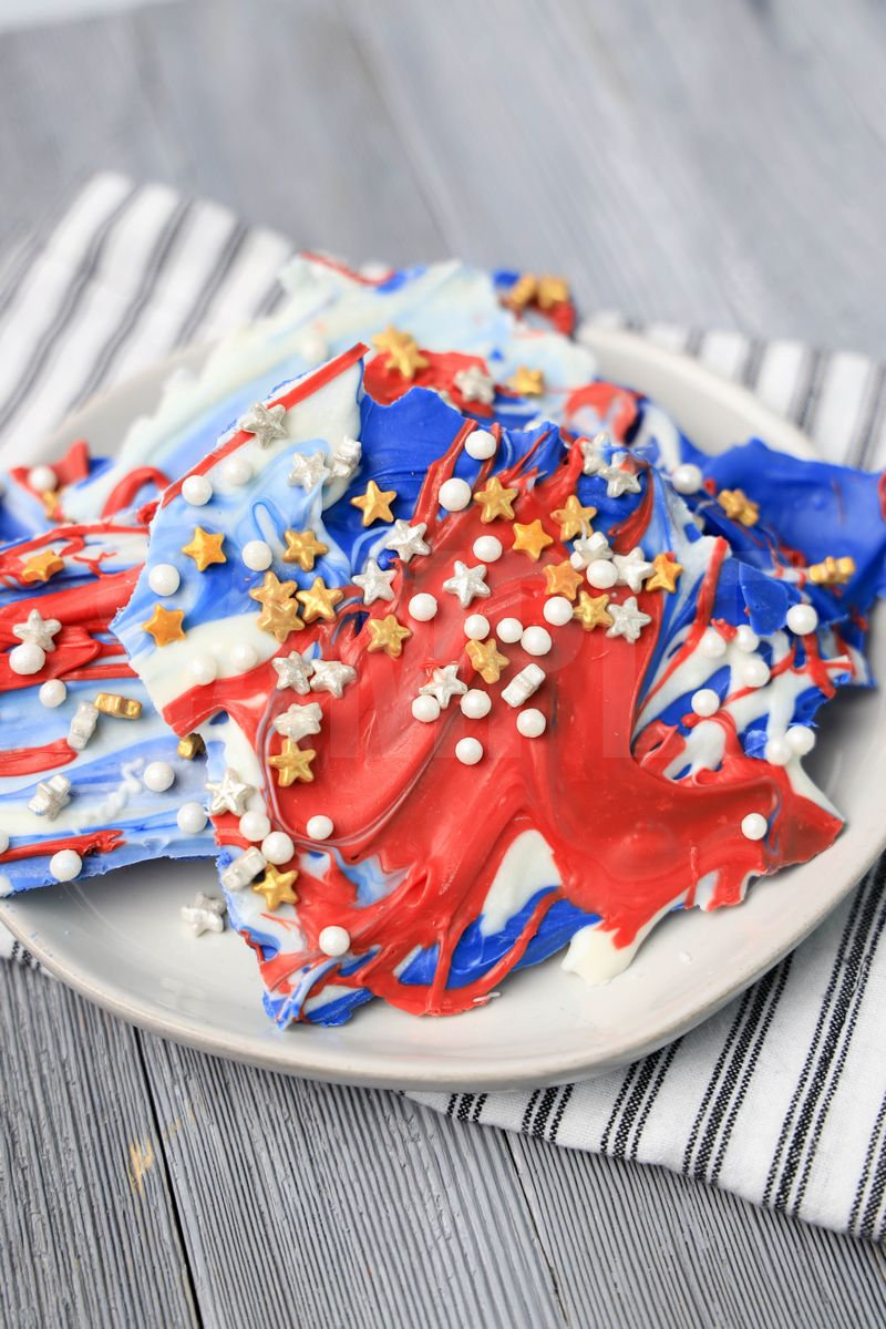 The 4th Of July Candy Bark comes on a white plate with a white striped napkin on a gray wood backdrop.