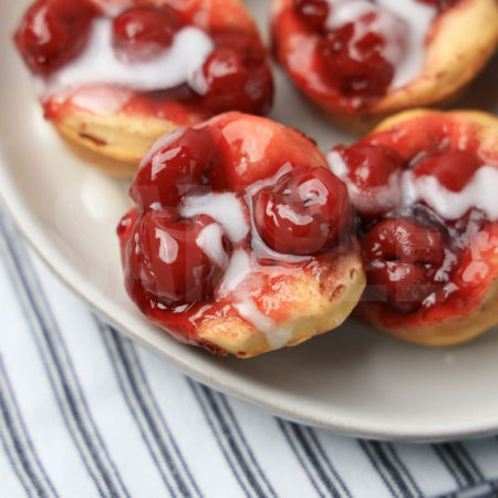 The Mini Cherry Cinnamon Roll Cups comes on a white plate with a white striped napkin on a gray wood backdrop.