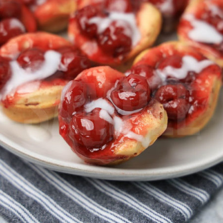 The Mini Cherry Cinnamon Roll Cups comes on a white plate with a gray striped napkin on a marble backdrop.