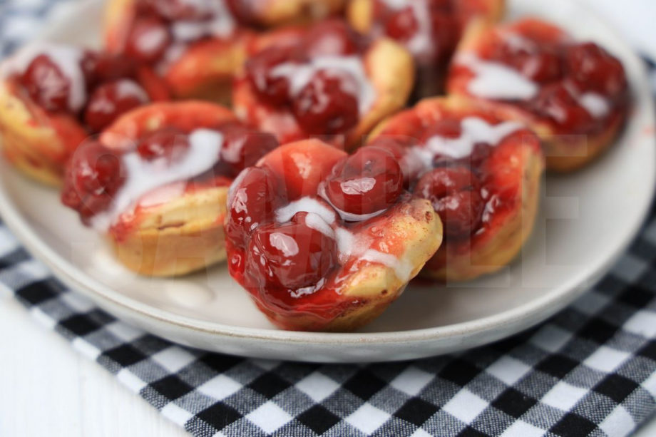 The Mini Cherry Cinnamon Roll Cups comes on a white plate with a plaid napkin on a white wood backdrop.