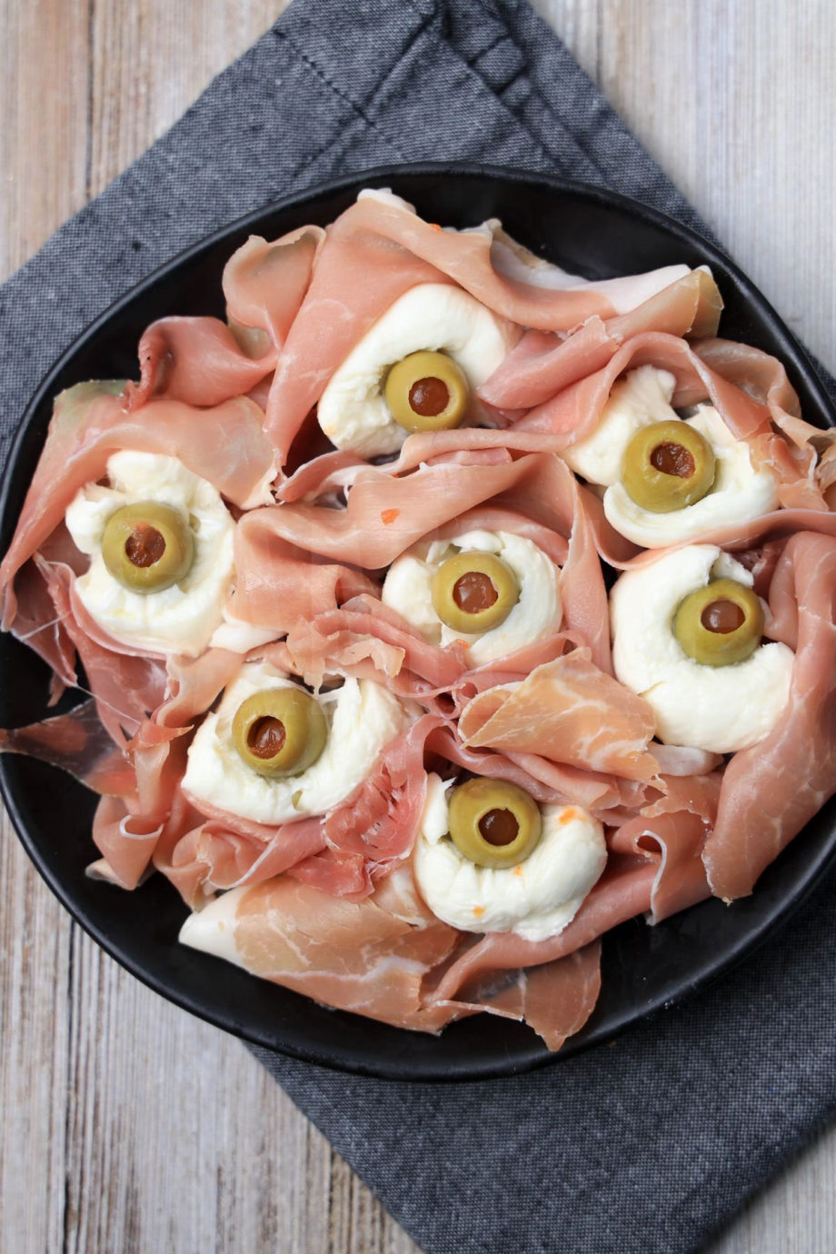The Prosciutto Eyeballs comes on a black plate with a denim napkin on a rustic wood backdrop.