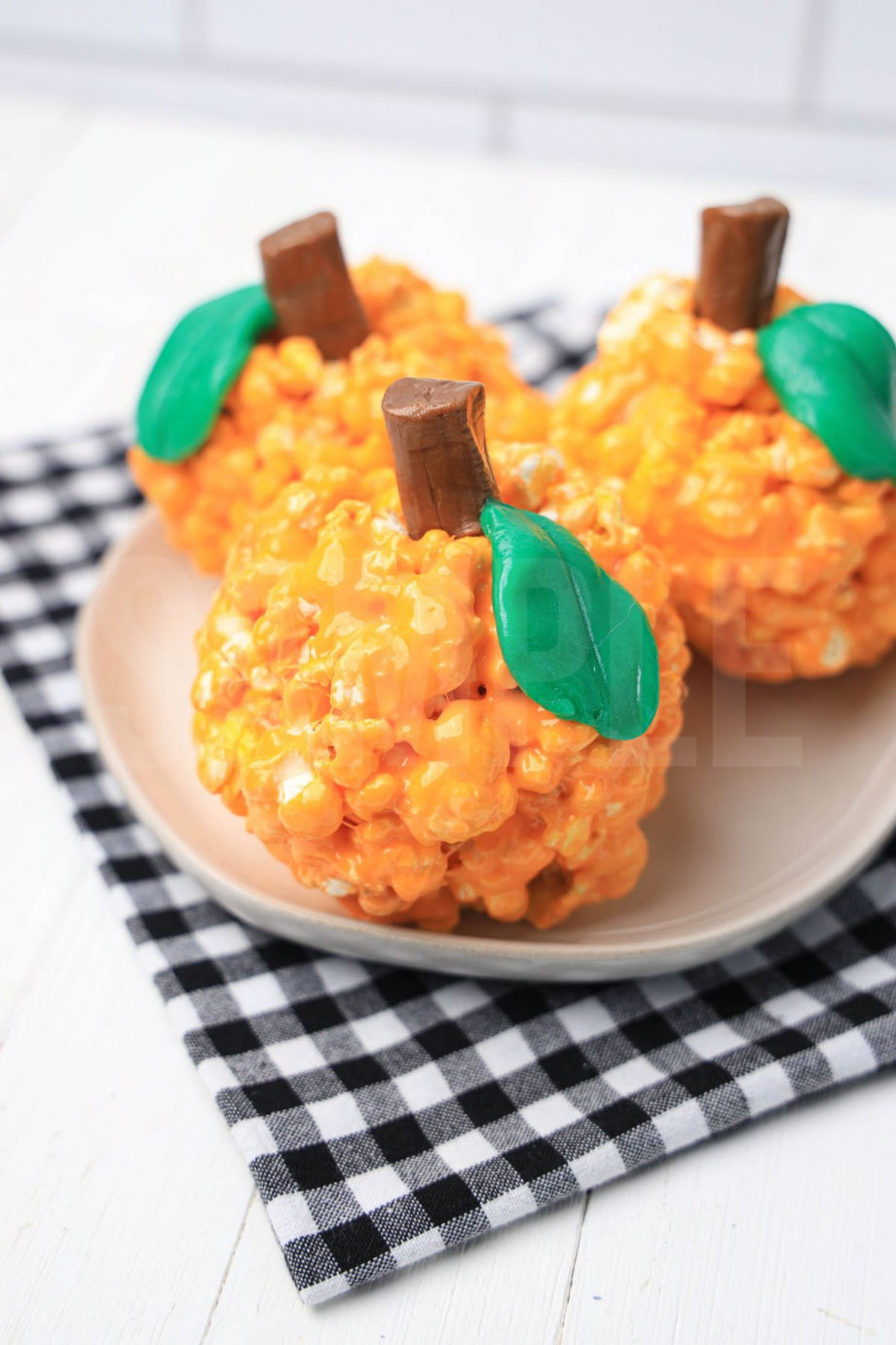 The Pumpkin Popcorn Balls comes on a white plate with a plaid napkin on a white wood backdrop.