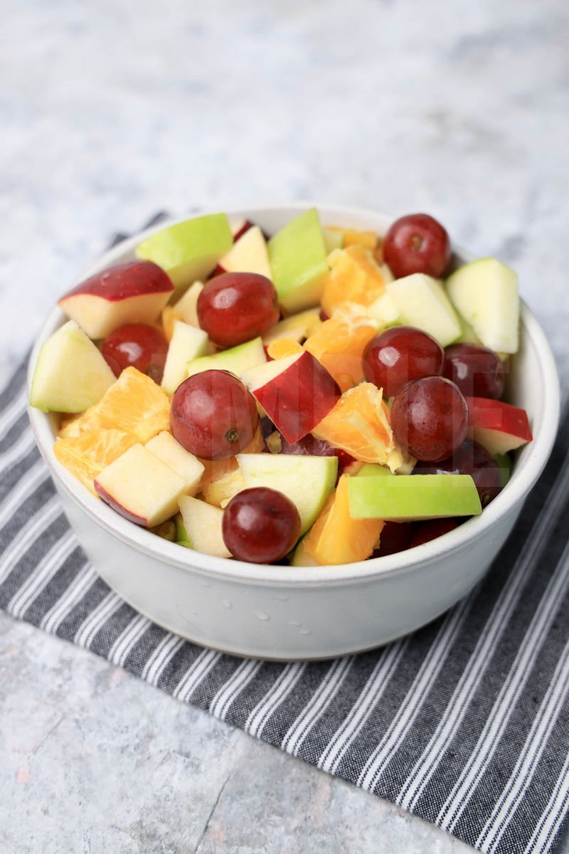 The Fall Fruit Salad comes in a white bowl with a gray striped napkin on a marble wood backdrop.