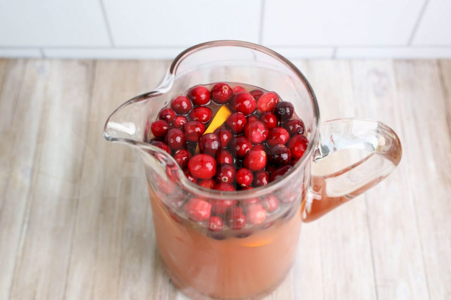 The Cranapple Holiday Punch comes in a clear glass pitcher on a rustic wood backdrop.