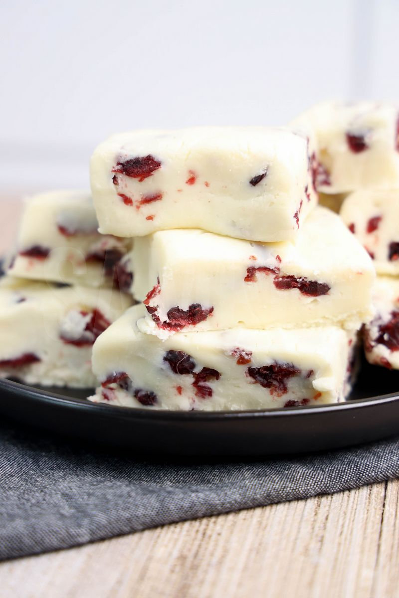 The White Chocolate Cranberry Fudge comes on a black plate with a denim napkin on a rustic wood backdrop.