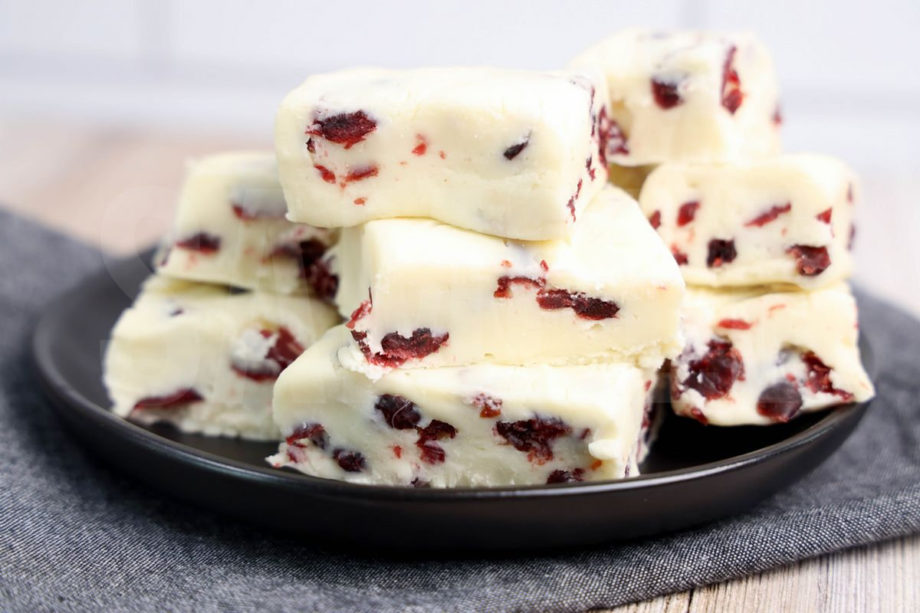 The White Chocolate Cranberry Fudge comes on a black plate with a denim napkin on a rustic wood backdrop.