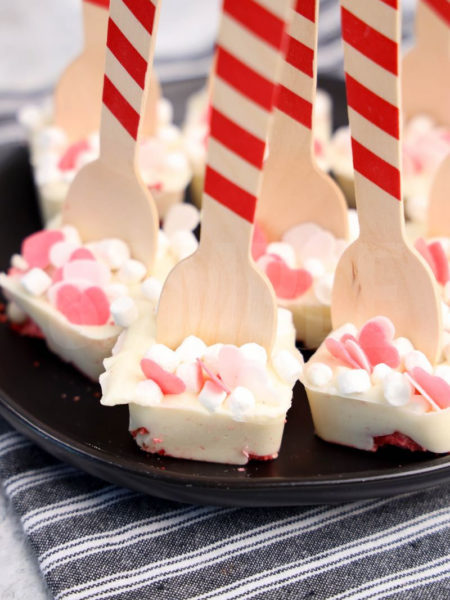 The Strawberry White Hot Chocolate Spoons comes on a black plate with a gray striped napkin on a marble wood backdrop.