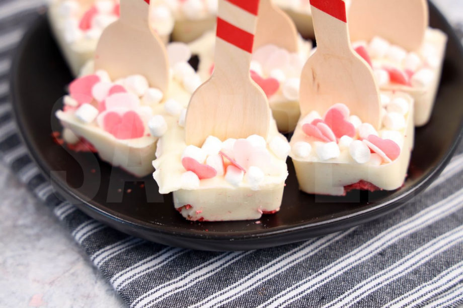 The Strawberry White Hot Chocolate Spoons comes on a black plate with a gray striped napkin on a marble wood backdrop.